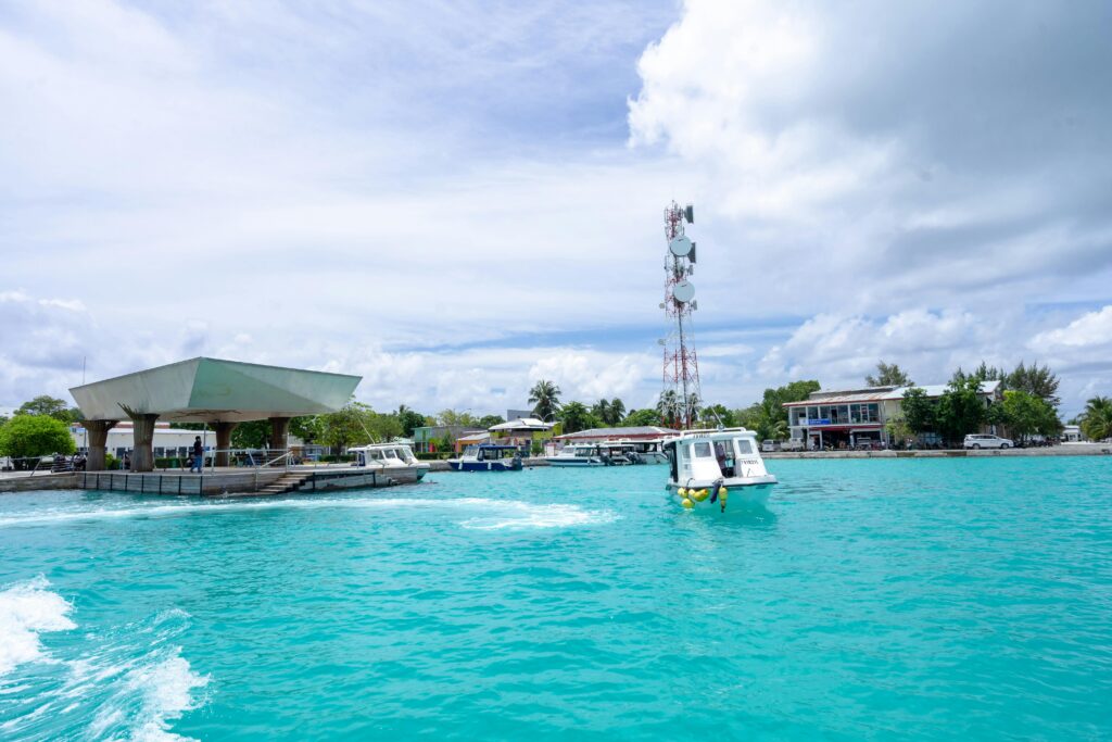 A tranquil tropical island scene featuring a dock, boats, and vibrant turquoise waters under a partly cloudy sky.